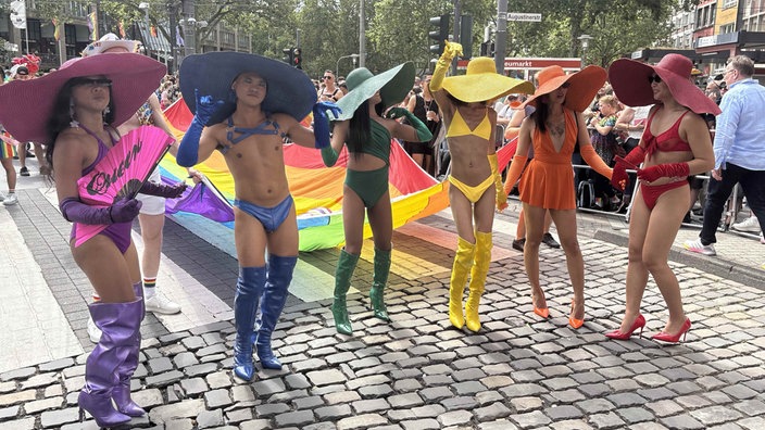 Sechs Personen in kurzem Kleid oder Bikini und großem Hut stehen im Paradenumzug. Jede trägt eine der Regenbogenfarben.