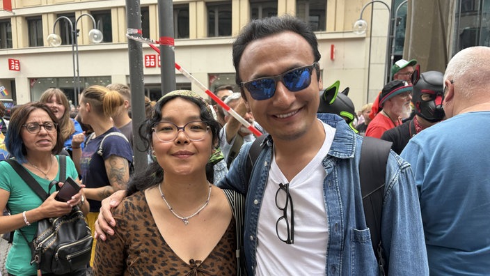 Ein Mann mit Sonnenbrille legt seinen Arm um eine neben íhm stehende Frau. Beide lächeln. 