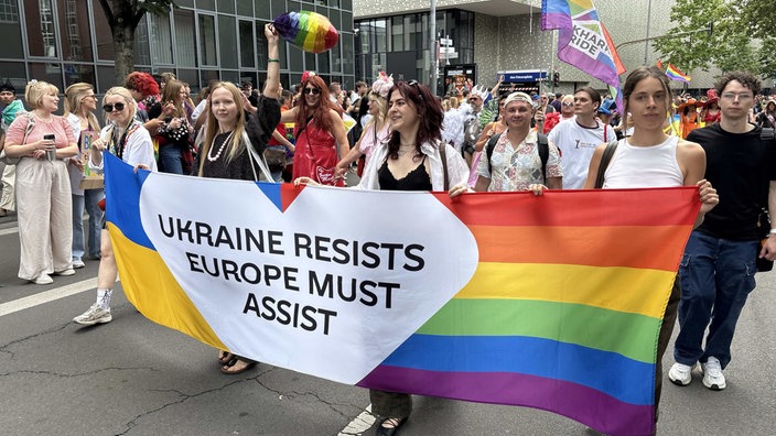 Vier Frauen tragen einen Banner, der die Farben der ukrainischen National- und der Prideflagge hat. In der Mitte steht auf einem weißen Herz mit schwarzer Schrift "Ukraine resists. Europe must assist" (Die Ukraine leistet Widerstand. Europa muss helfen).