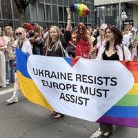 Vier Frauen tragen einen Banner, der die Farben der ukrainischen National- und der Prideflagge hat. In der Mitte steht auf einem weißen Herz mit schwarzer Schrift "Ukraine resists. Europe must assist" (Die Ukraine leistet Widerstand. Europa muss helfen).