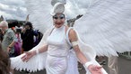 Eine Drag-Queen in weißem Glitzer-Kleid und Engelsflügel lächelt in die Kamera.
