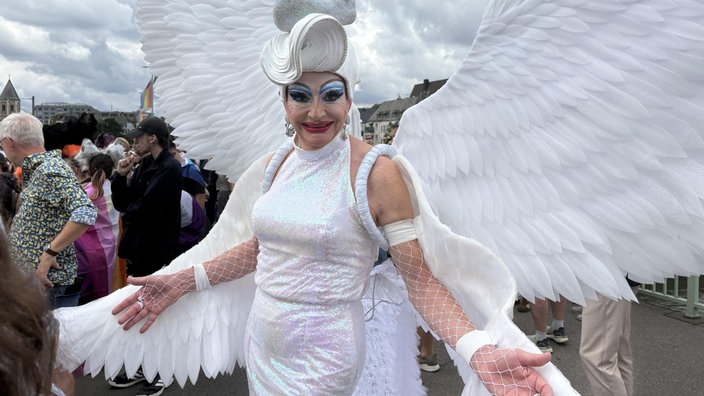 Eine Drag-Queen in weißem Glitzer-Kleid und Engelsflügel lächelt in die Kamera.