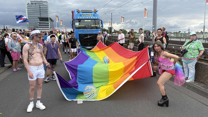 Teilnehmer des CSD tragen eine meterlange Regenbogenflagge vor einem blauen Lkw her.