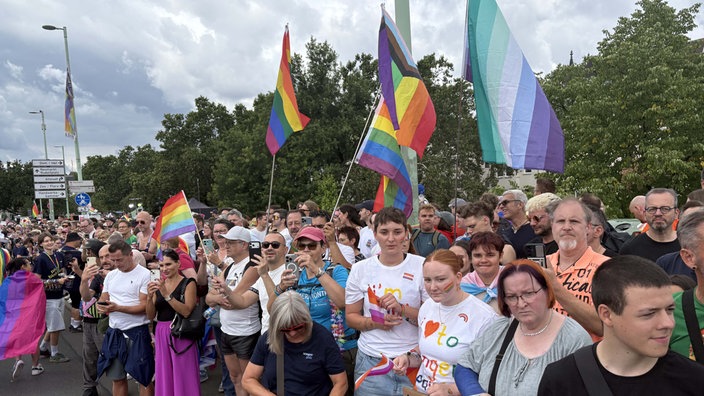 Am Straßenrand stehen Menschen, die Regenbogen-Flaggen schwenken oder mit ihren Smartphones filmen.  