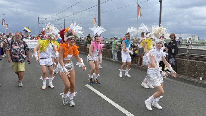Weißgekleidete Tänzerinnen und Tänzer mit buntem Federschmuck performen auf einer Rheinbrücke.