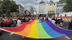 Köln Heumarkt: Teilnehmer des CSD 2026 in Köln tragen eine gigantische Regenbogenflagge. Ringsum erstreckt sich eine Menschenmasse.