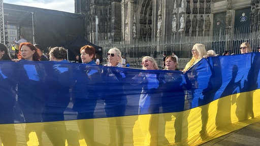 Mehrere Personen halten einen Banner mit der Flagge der Ukraine hoch und stehen vor dem Kölner Dom.