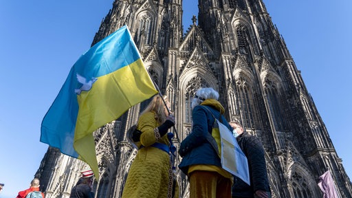 Drei Personen stehen vor dem Kölner Dom. Eine von ihnen hält eine Flagge der Ukraine.