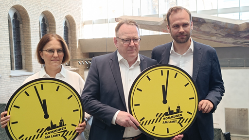 Kölns Oberbürgemeister, die städtische Kämmerin und der Schuldezernent stehen nebeneinander und halten zwei gelbe große Uhren aus Pappe hoch, wo draufsteht Kommunen am Limit.