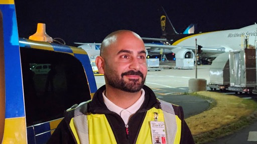 Can Talay in Warnweste vor einem Flugzeug auf dem nächtlich beleuchteten Gelände des Flughafens Köln/Bonn.