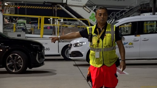 Yalcin Alibas in Warnweste und auffälliger Arbeitskleidung weist auf dem Gelände des Flughafens Köln/Bonn mit ausgestrecktem Arm die Richtung.