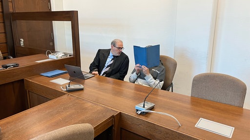 Ein Mann verdeckt sein Gesicht mit einem Ordner, daneben sitzt ein Mann mit Anzug