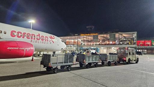 Gepäckwagen werden nachts neben einem Flugzeug vor dem beleuchteten Terminal des Flughafens Köln/Bonn transportiert.