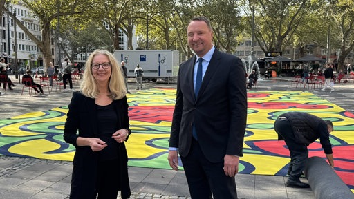 Drei Personen stehen auf dem Kölner Neumarkt. Im Hintergrund ist ein großer bunter Teppich zu sehen.