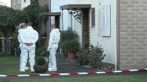 Zwei Männer und eine Frau in weißen Schutzanzügen stehen in einem Vorgarten vor einem beigen Haus. Vor und hinter ihnen ist rot-weißes Absperrband gespannt.