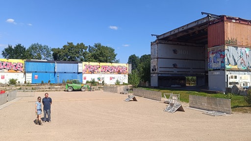 Ein leeres Festivalgelände. Die Bühne besteht aus drei gestapelten Containern auf jeder Seite. Links unten im Bild stehen ein Mann und eine Frau, die in die Kamera schauen.