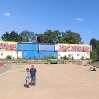 Ein leeres Festivalgelände. Die Bühne besteht aus drei gestapelten Containern auf jeder Seite. Links unten im Bild stehen ein Mann und eine Frau, die in die Kamera schauen.