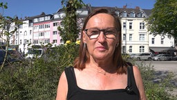 Eine Frau mit braunen schulterlangen Haaren und einer Brille schaut ernst in die Kamera. Im Hintergrund ist eine Häuserfassade.
