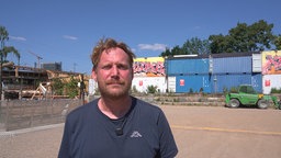 Ein Mann mit roten Haaren schaut ernst in die Kamera. Im Hintergrund sind Container.
