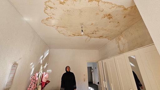 Eine Frau steht in einem Zimmer und guckt sich die Zimmerdecke an, auf der sich Schimmel breit gemacht hat