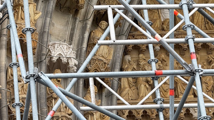 Hinter einem Gerüst erkennt man verschiedene Figuren und einen hellen Baldachin.