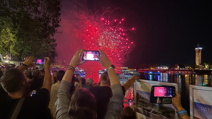 Es wird von hinten auf mehrere Menschen gefilmt, die auf den Rhein schauen, auf dem gerade ein pinkes Feuerwerk leuchtet. Die Leute heben die Handys und filmen. 