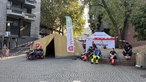 Vor einer Erste-Hilfe-Station der Malteser sitzen Sanitäter auf einer Bank.