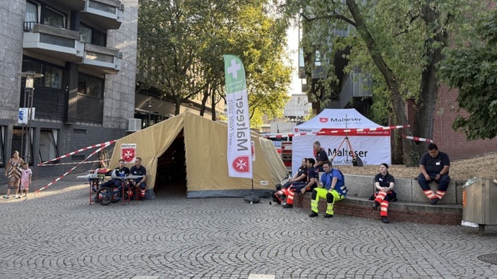 Vor einer Erste-Hilfe-Station der Malteser sitzen Sanitäter auf einer Bank.