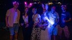 Mehrere Menschen stehen im Dunkeln nebeneinander und tragen verschiedene Leuchtaccessoires