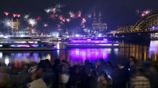 zwei Schiffe fahren bei Nacht auf dem Rhein. Im Vordergrund eine Menschenmenge; im Hintergrund der Kölner Dom und Feuerwerk im Nachthimmel.