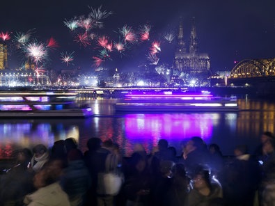 zwei Schiffe fahren bei Nacht auf dem Rhein. Im Vordergrund eine Menschenmenge; im Hintergrund der Kölner Dom und Feuerwerk im Nachthimmel.
