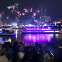 zwei Schiffe fahren bei Nacht auf dem Rhein. Im Vordergrund eine Menschenmenge; im Hintergrund der Kölner Dom und Feuerwerk im Nachthimmel.