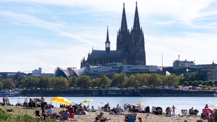 Am Rheinufer sitzen Menschen mit Sonnenschirmen und Picknickdecken, im Hintergrund der Dom.