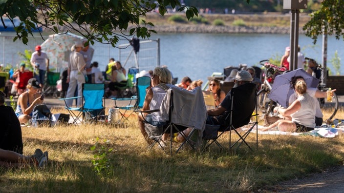 Eine Menschengruppe sitzt auf Campingstühlen auf einer Wiese am Rheinufer.