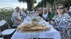 Eine Gruppe von Menschen sitzt an einem langen gedeckten Tisch am Rheinufer.