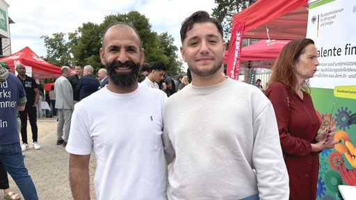 Roni Özkul (Sohn) und Gökmen Özkul (Vater) stehen auf einer Ausbildungsmesse neben einem roten Zelt