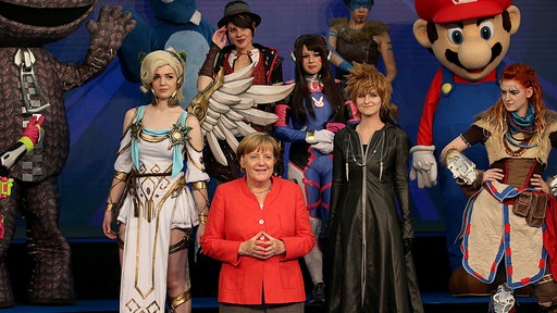 Angela Merkel in rotem Blazer steht zwischen jungen Menschen, die als Figuren aus Computerspielen verkleidet sind