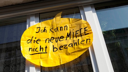 Vor einem Fenster eines Mehrfamilienhauses ist ein gelbes Plakat angebracht. Darauf steht: "Ich kann die neue Miete nicht bezahlen"