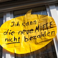Vor einem Fenster eines Mehrfamilienhauses ist ein gelbes Plakat angebracht. Darauf steht: "Ich kann die neue Miete nicht bezahlen"