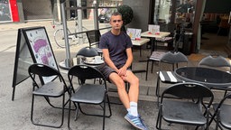 Niklas Kaiser von ColognePride sitzt in einem Kölner Café und schaut in die Kamera: