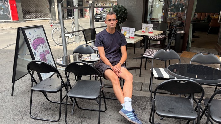 Niklas Kaiser von ColognePride sitzt in einem Kölner Café und schaut in die Kamera: