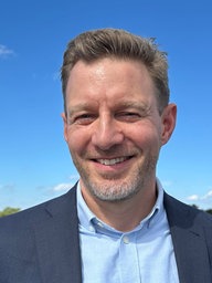 Nils Müller, ein Mann in Anzug, steht auf einem Dach. Im Hintergrund blauer Himmel.