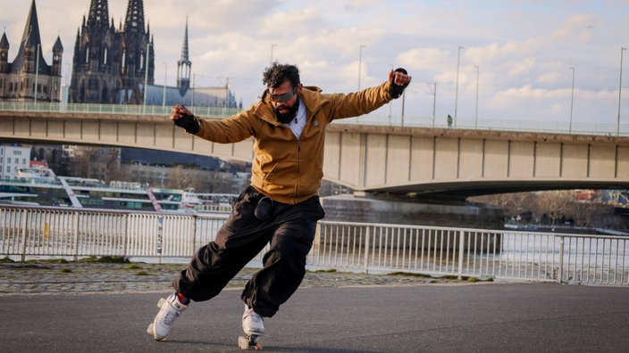 Ein Mann (Pedro Corona) macht auf Inline-Skatern eine Kunstfigur. Im Hintergrund der Kölner Dom