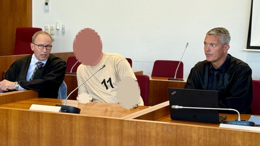 Der Angeklagte sitzt in der Mitte und wurde unkenntlich gemacht. Rechts und links von ihm sitzt seine Rechtsberatung.