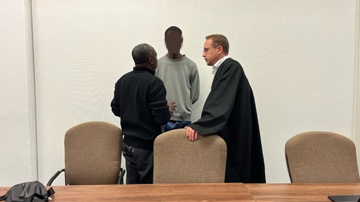 Der Angeklagte in dem Prozess steht mit seinem Dolmetscher (links) und seinem Verteidiger (rechts) im Gerichtssaal am Landgericht Köln