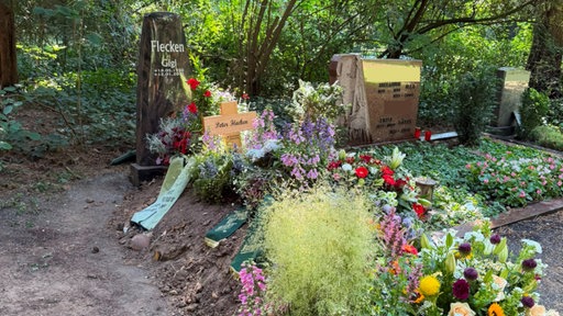 Ein frisch ausgehobenes Grab, das mit Blumen und Kränzen bedeckt ist, an der Seite liegt ein Schädelfragment.