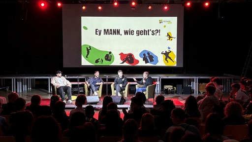 Vier Männer auf einem Podium. Darüber Bildschirm mit der Aufschrift: "Ey, MANN, wie geht's?!"
