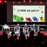 Vier Männer auf einem Podium. Darüber Bildschirm mit der Aufschrift: "Ey, MANN, wie geht's?!"