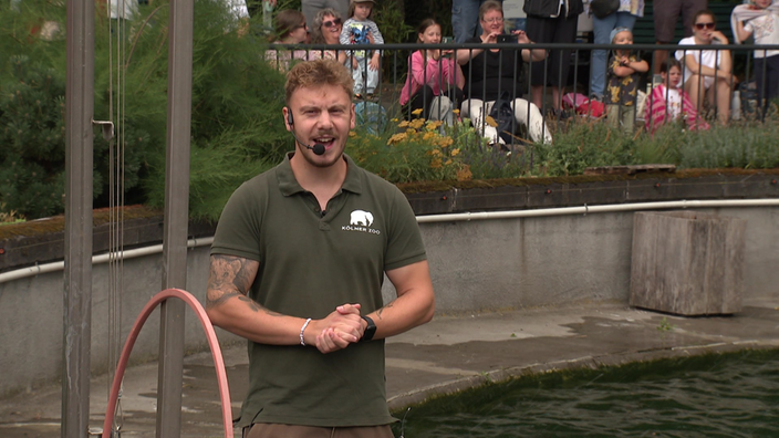 Tierpfleger Tim Burghof mit Head-Set Mikrofon und im Hintergund viele Zoobesucher.