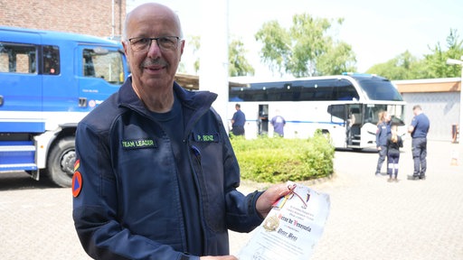 Ein Mann mit weißen Haaren und Brille in blauer THW-Kleidung hält eine Urkunde mit der Aufschrift "Héroe de Venezuela".